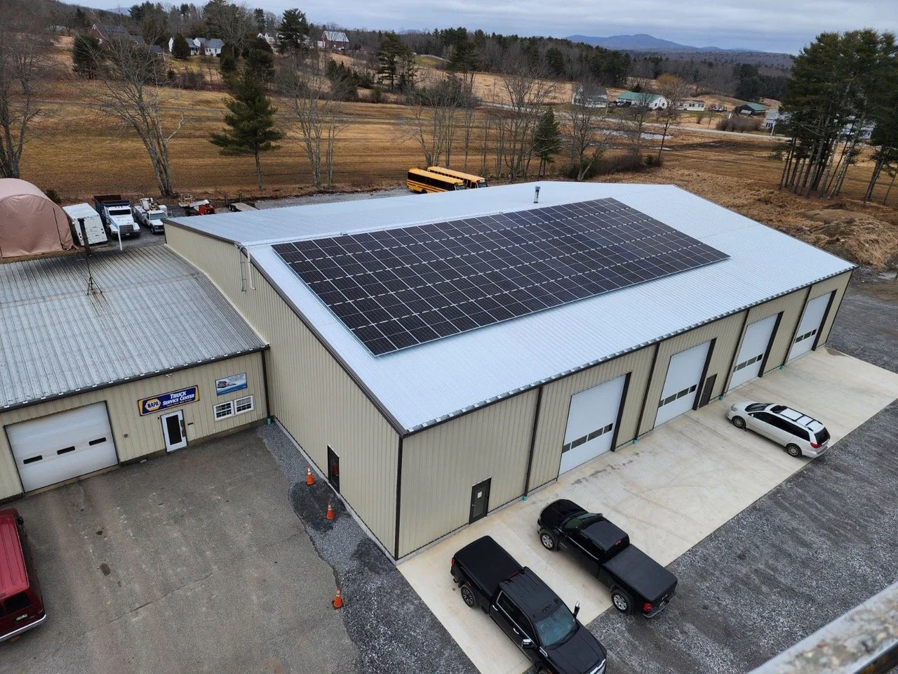 Large industrial building with solar panels on the roof, several garage doors, parked cars and trucks, and rural landscape in the background.