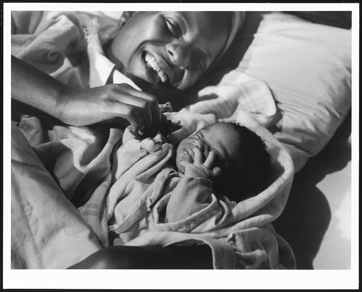 A woman and a baby lying on a bed, smiling and playing together in black and white. The woman appears joyful as she interacts with the baby.