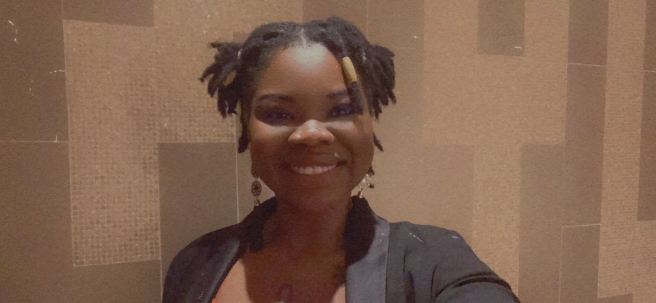 A woman taking a selfie in front of a tiled wall, smiling, wearing earrings and a dark blazer, with short dreadlocks styled with beige hair beads.