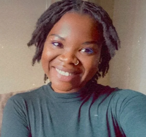 Smiling woman with short dreadlocks, nose ring, and dark shirt in indoor setting.