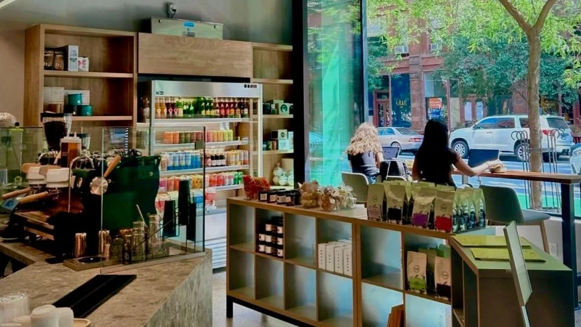 Inside a coffee shop showing a counter with coffee equipment, shelves with jars and products, a refrigerator with drinks, and two women sitting near large windows looking outside at parked cars and trees.