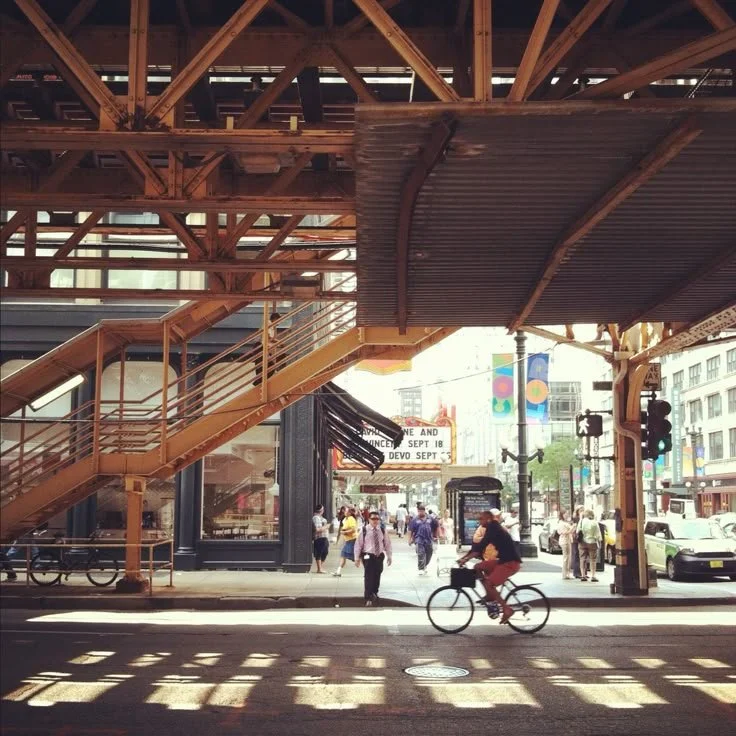 Street view with people walking and riding bikes underneath an elevated train platform with metal stairs and support beams.