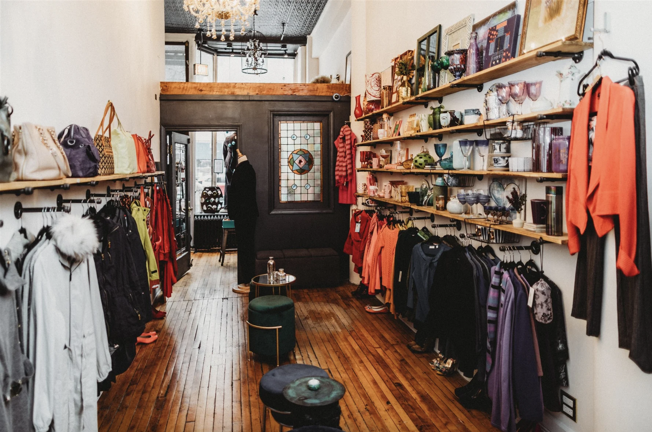Inside a boutique clothing store with shelves of colorful clothing, handbags, and decorative glassware, featuring wooden floors, a black wall with stained glass window, and a mannequin near the entrance.
