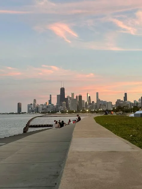 People sitting on a curved pier along a lake at sunset with the Chicago skyline in the background.