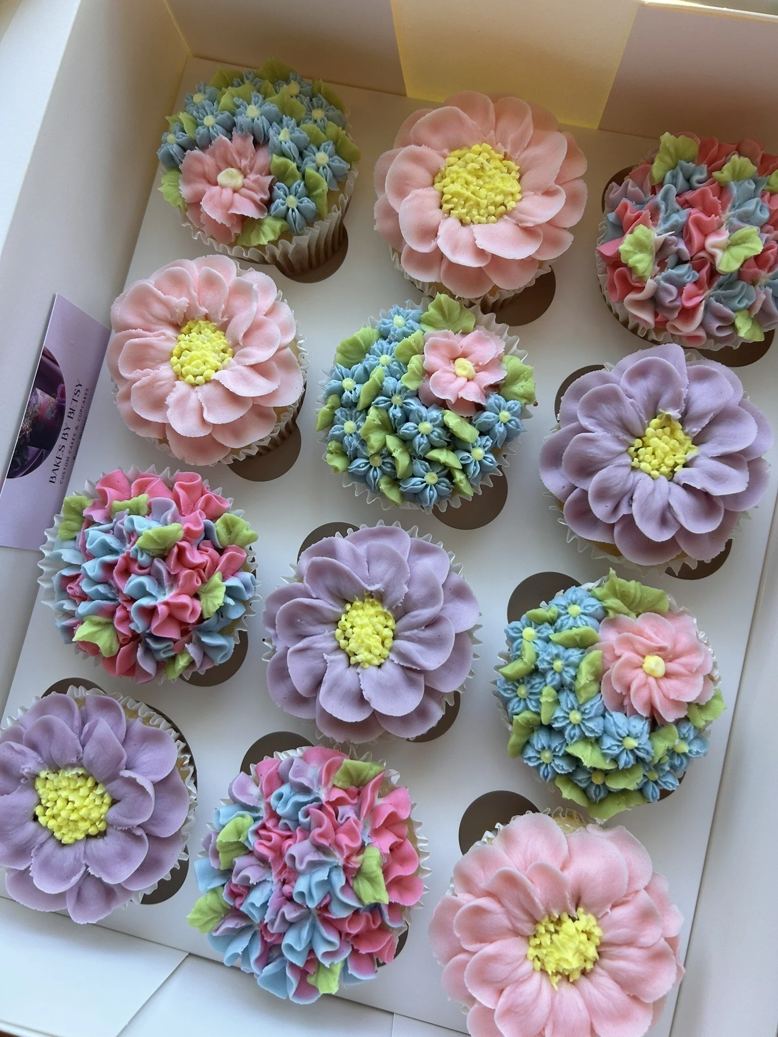 A collection of cupcake pastries decorated with white and green icing in flower and floral patterns, arranged in white boxes.