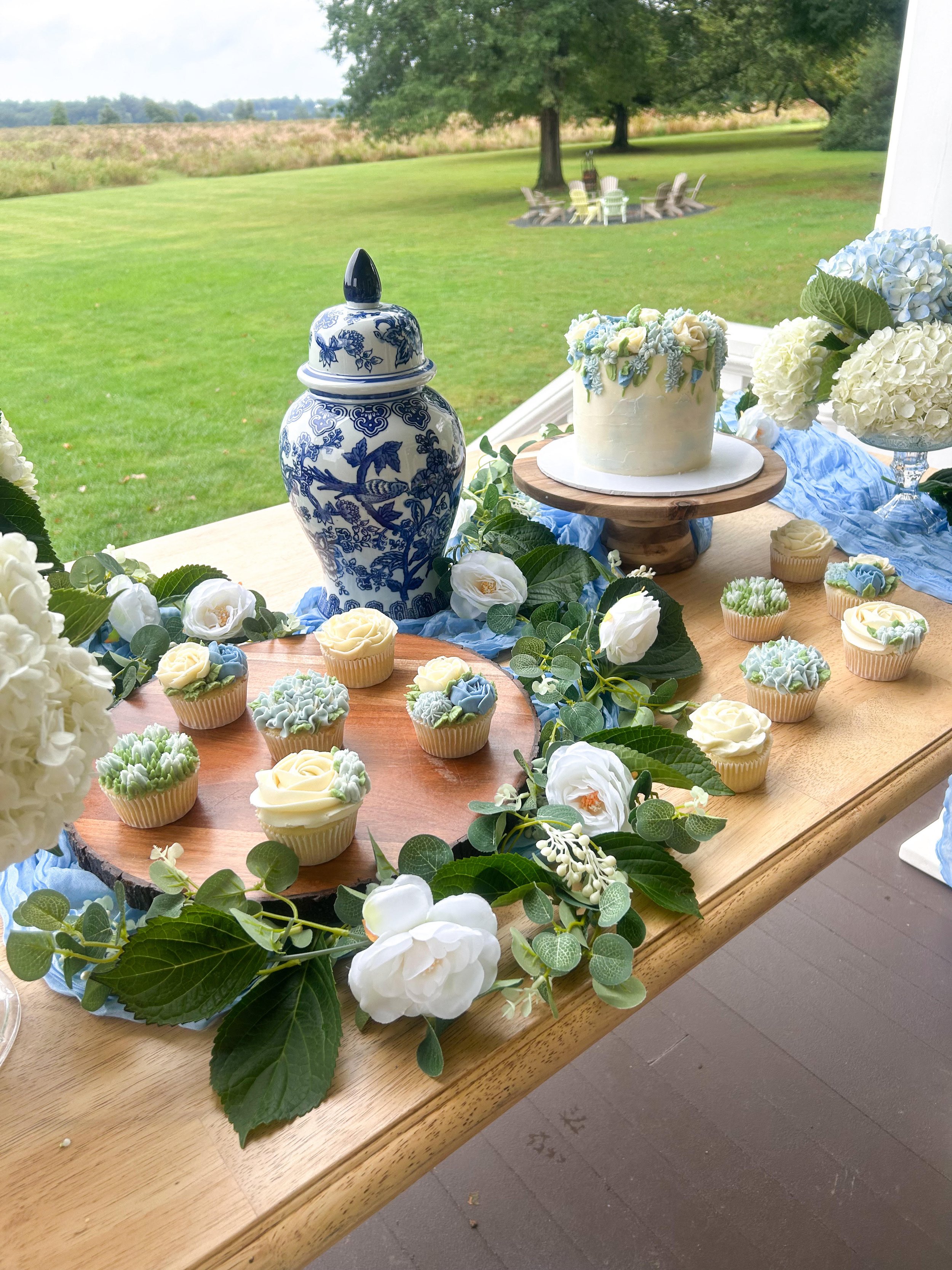 Decorated table outdoors with cupcakes, a cake, floral arrangements, and a blue and white vase, overlooking a grassy yard with trees.