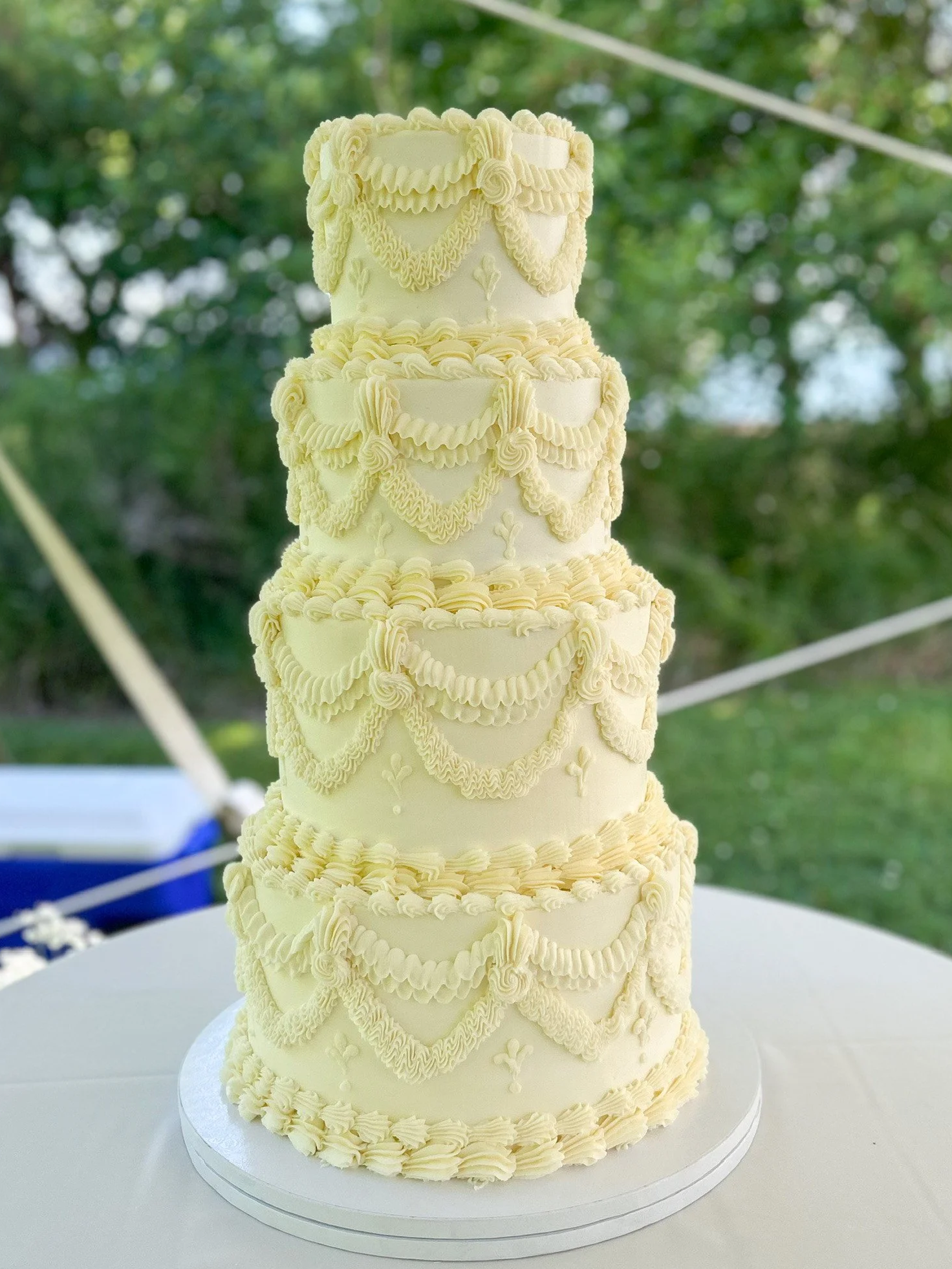 Four-tiered wedding cake with white frosting, red ribbon bands, yellow sunflowers, and purple and white flowers, placed on a wooden log slice