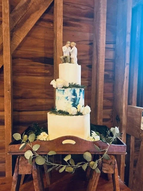 Three-tiered wedding cake with a bride and groom figurine topper, decorated with white flowers and greenery, placed on a wooden stand inside a rustic barn.