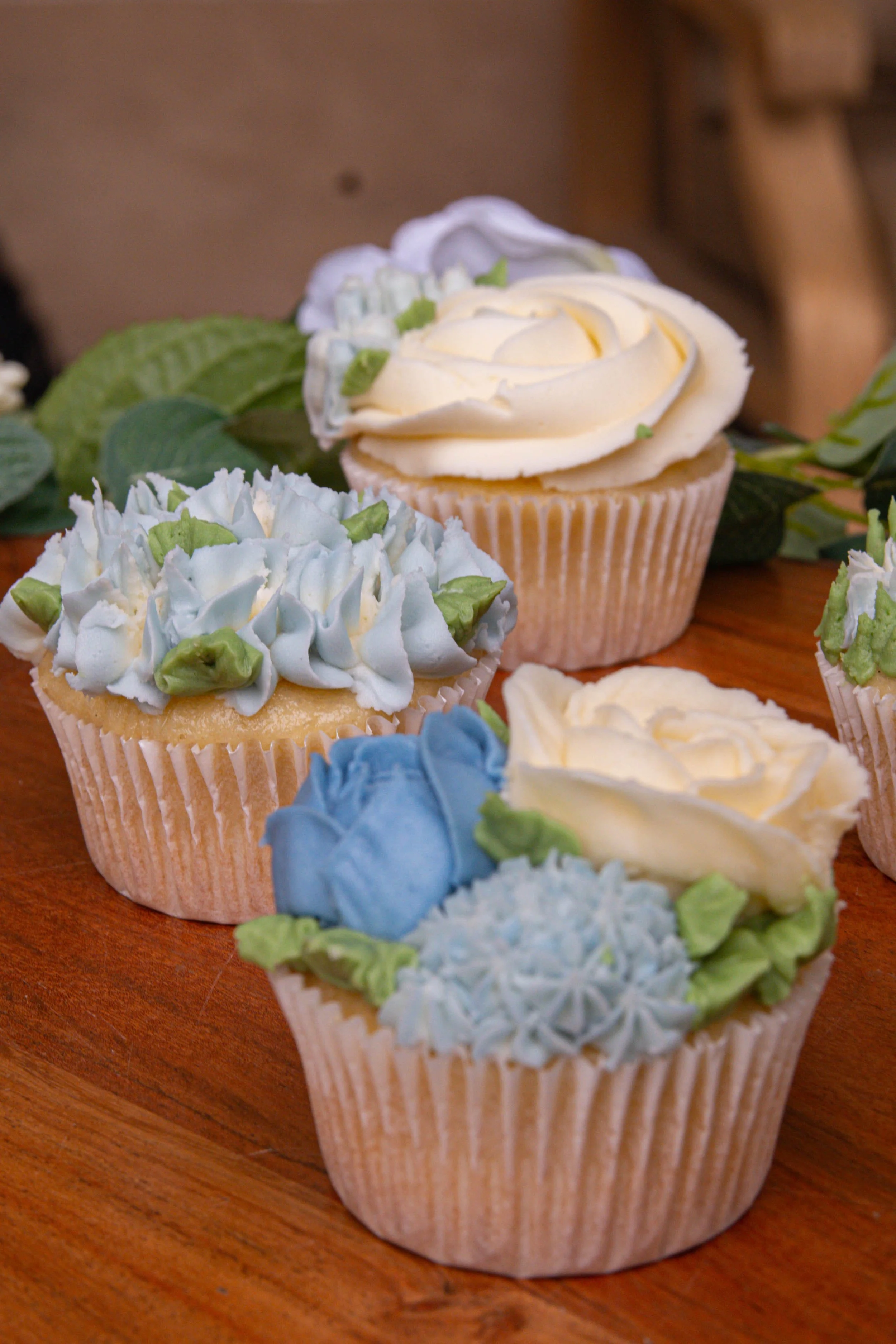 Decorated cupcakes with white, blue, and green frosting and floral decorations on a wooden surface.