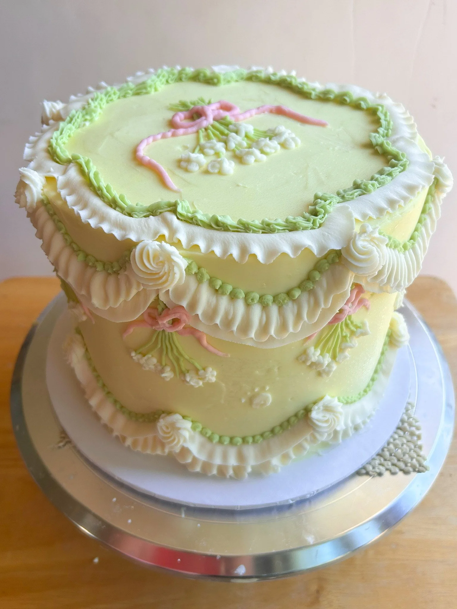 A cake with white frosting and yellow flowers, decorated with green leaves, and a message that says 'God Bless Anna' in orange and pink lettering. The cake is on a glass stand.