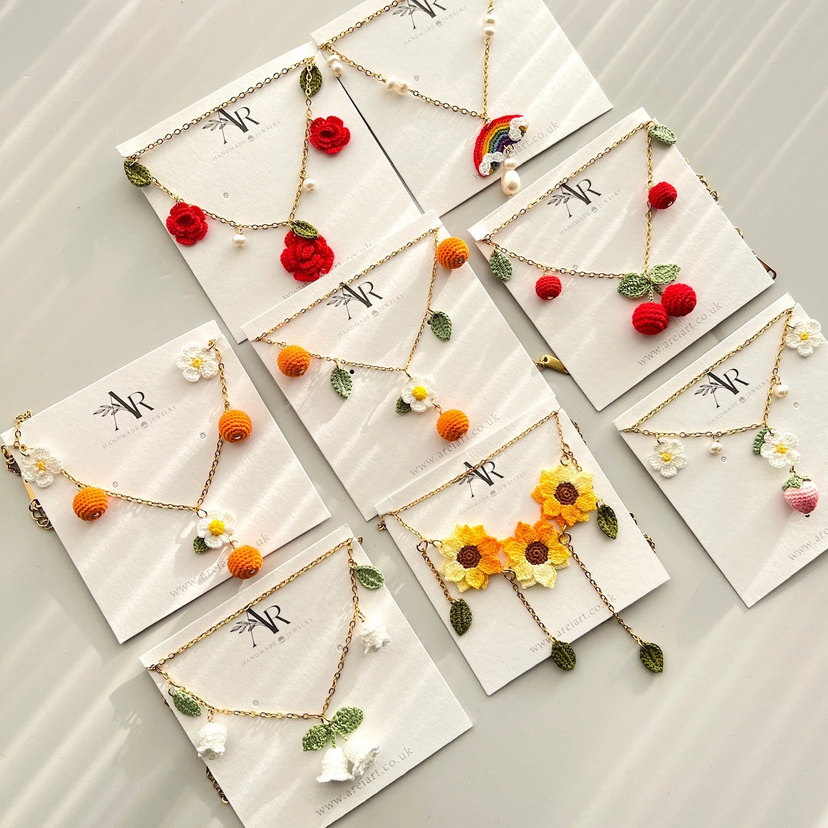 Display of six handmade jewelry necklaces with crocheted floral and fruit charms on branded white cards.