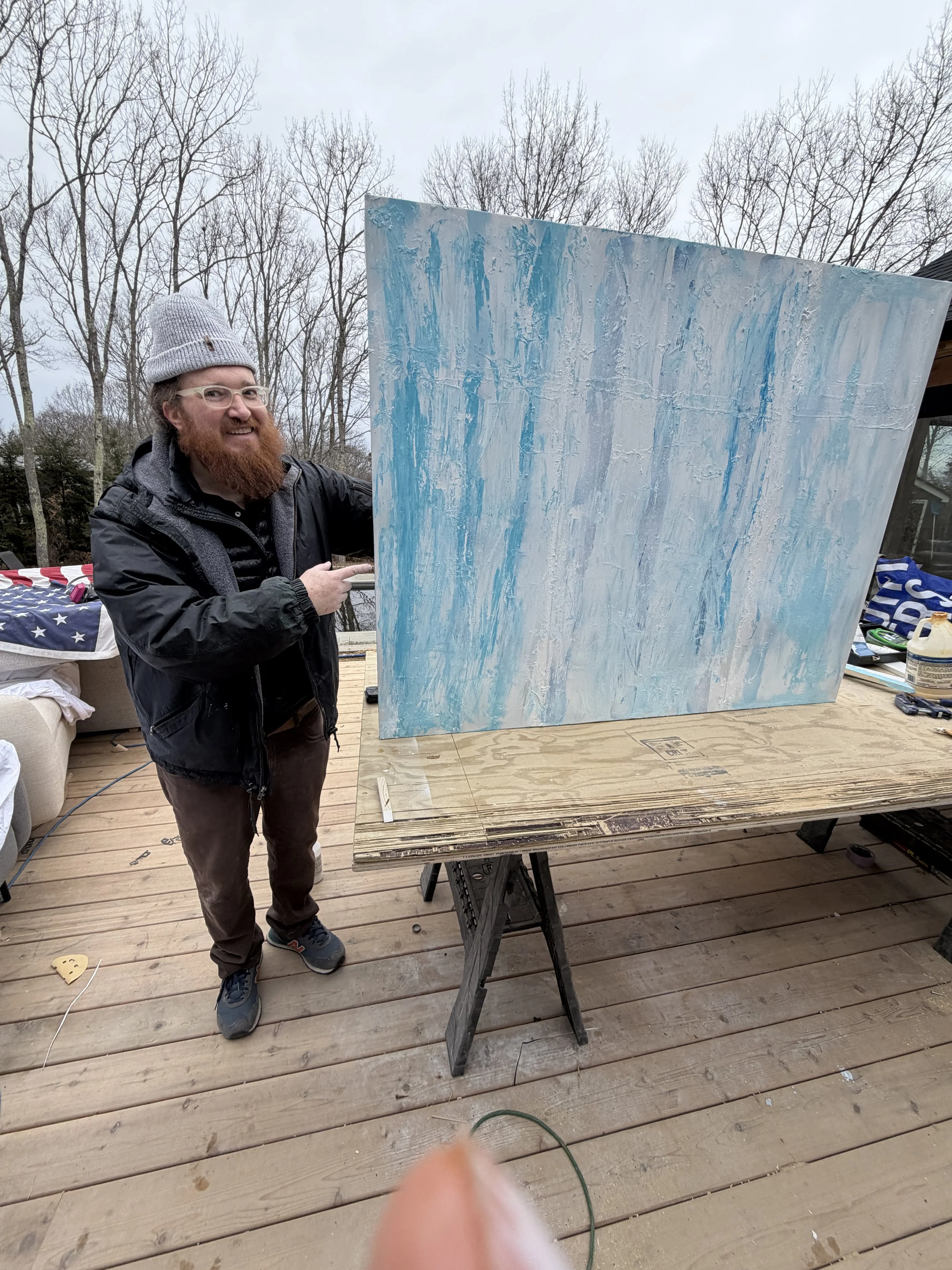 A man with a red beard, glasses, a gray knit hat, and a black jacket is smiling and pointing at a large abstract painting with blue and white textured brushstrokes on an easel outdoors on a wooden deck. Bare trees are in the background.