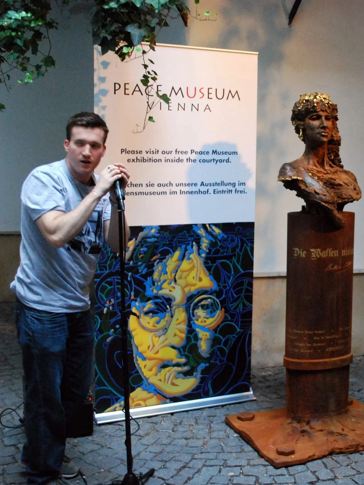 Young man holding microphone at the Peace Museum Vienna, standing beside a classical bust sculpture on a pedestal.