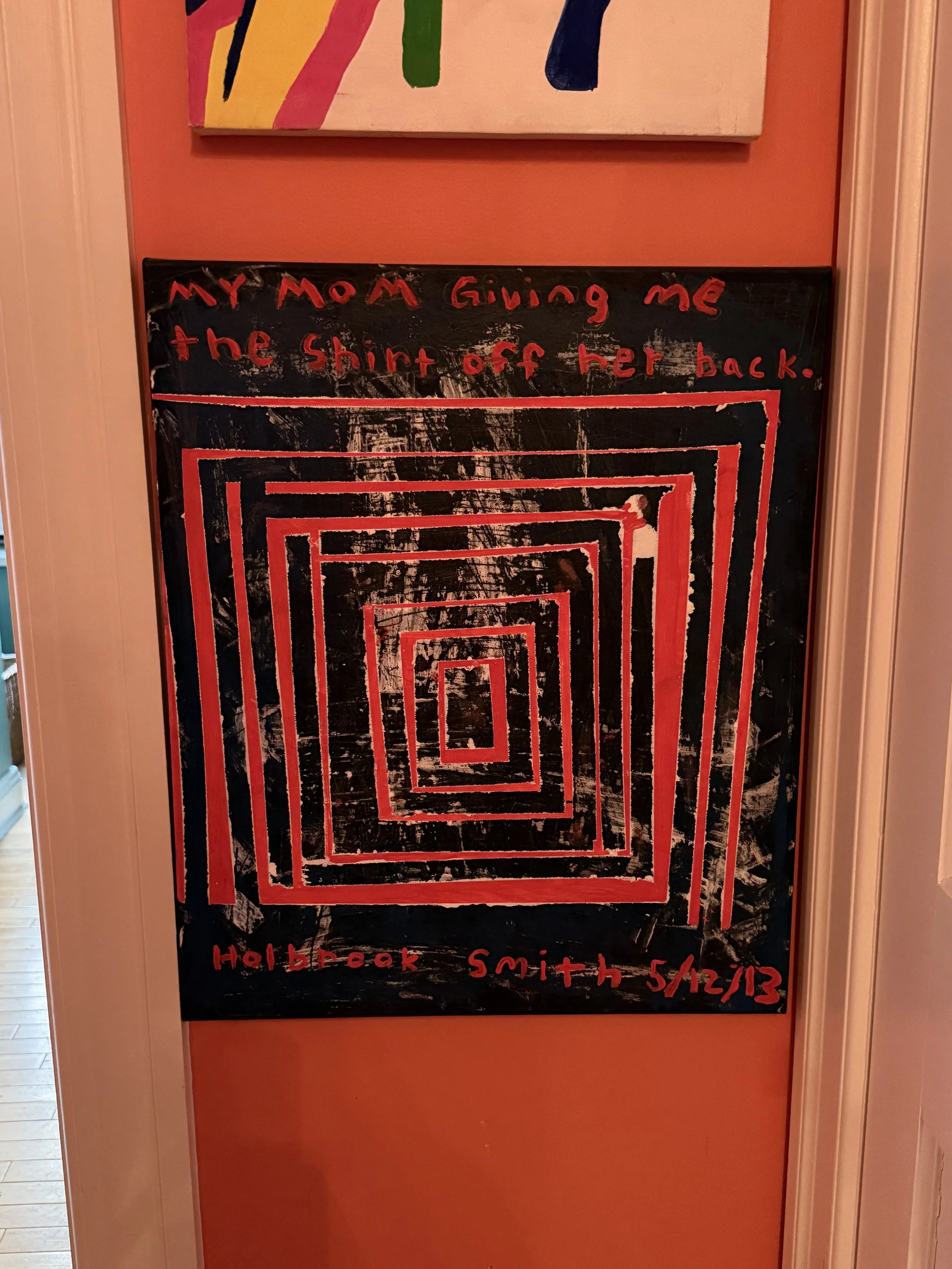 A blackboard with red, hand-written text that reads 'My mom giving me the shirt off her back.' and a spiral of red square outlines. At the bottom, it says 'Hollbrook Smith 5/22/13'. The background wall is orange, and there's an abstract colorful pain