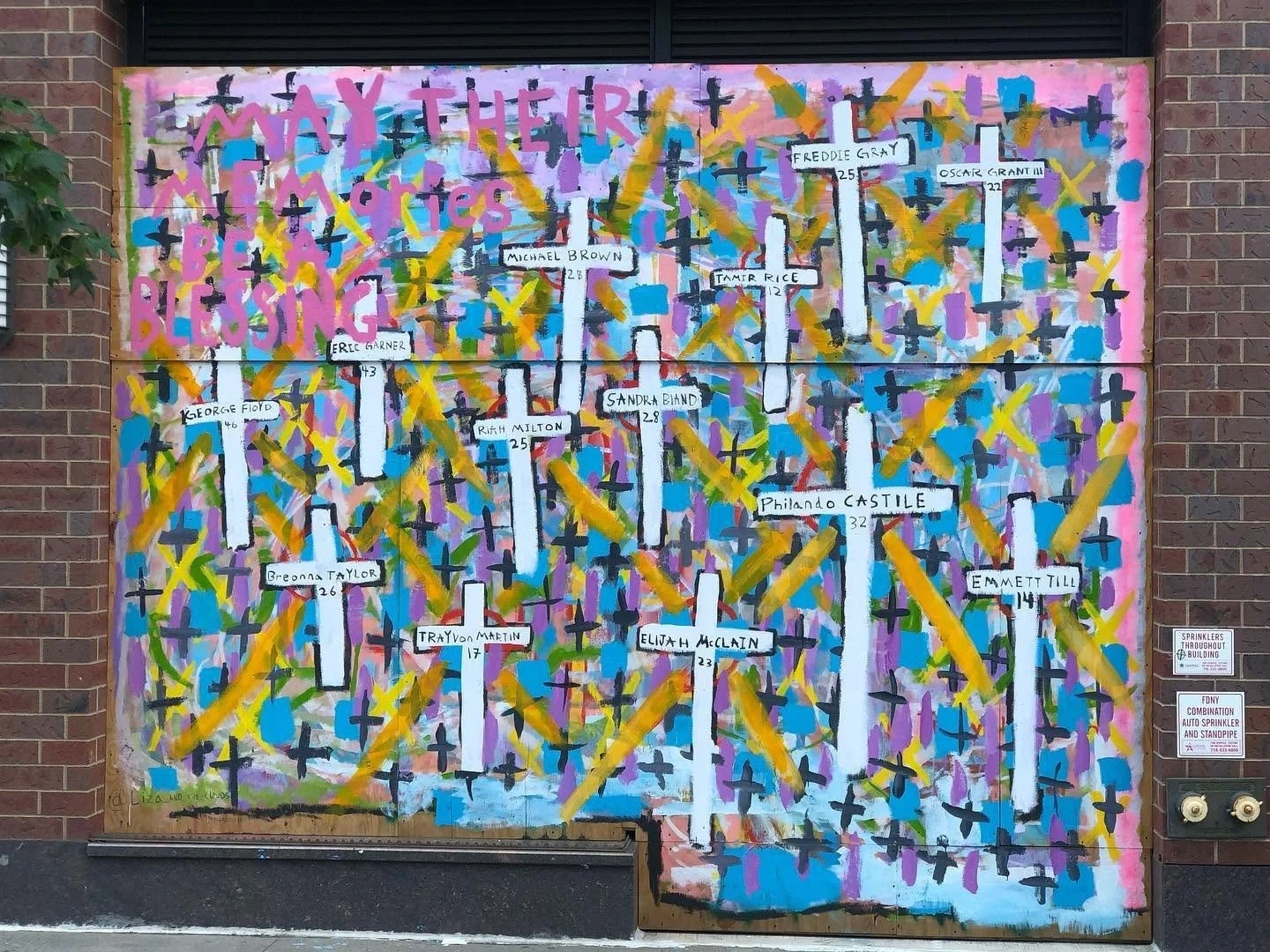 Colorful mural with crosses, yellow strokes, pink background, and names of victims of violence, with some names and their ages written on white vertical bars.