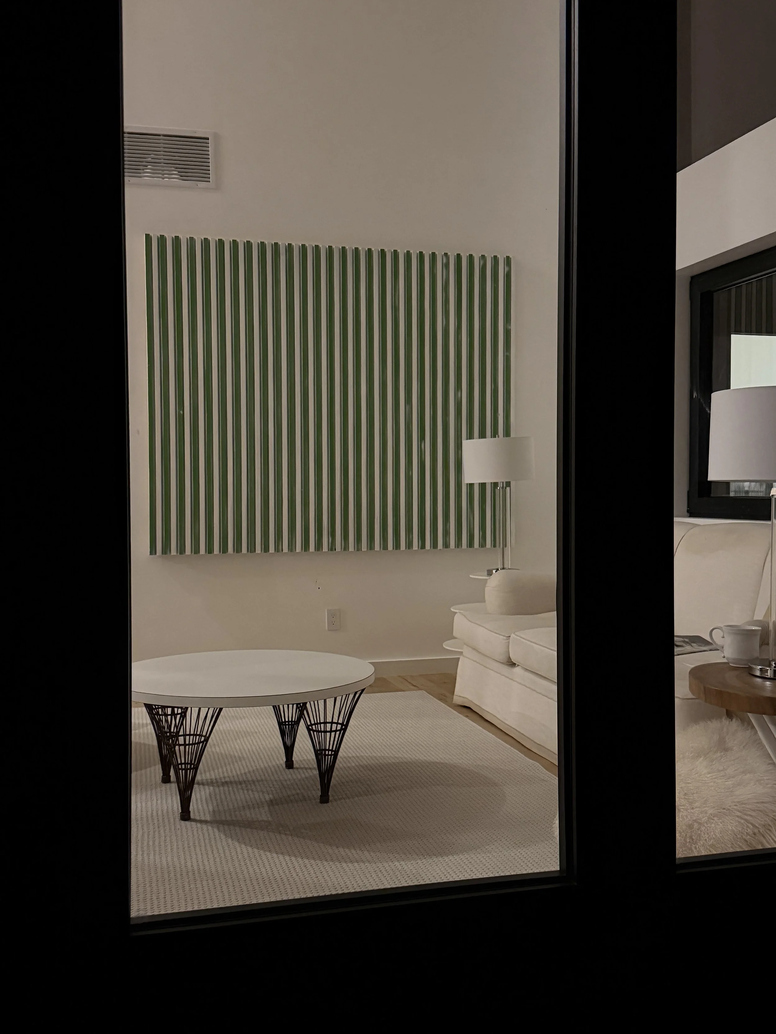 Interior view of a modern living room seen through a glass door, featuring a white sofa with a lamp behind it, a wooden side table with a teacup and a magazine, a round coffee table with a wireframe design, green and white striped wall decor, a white