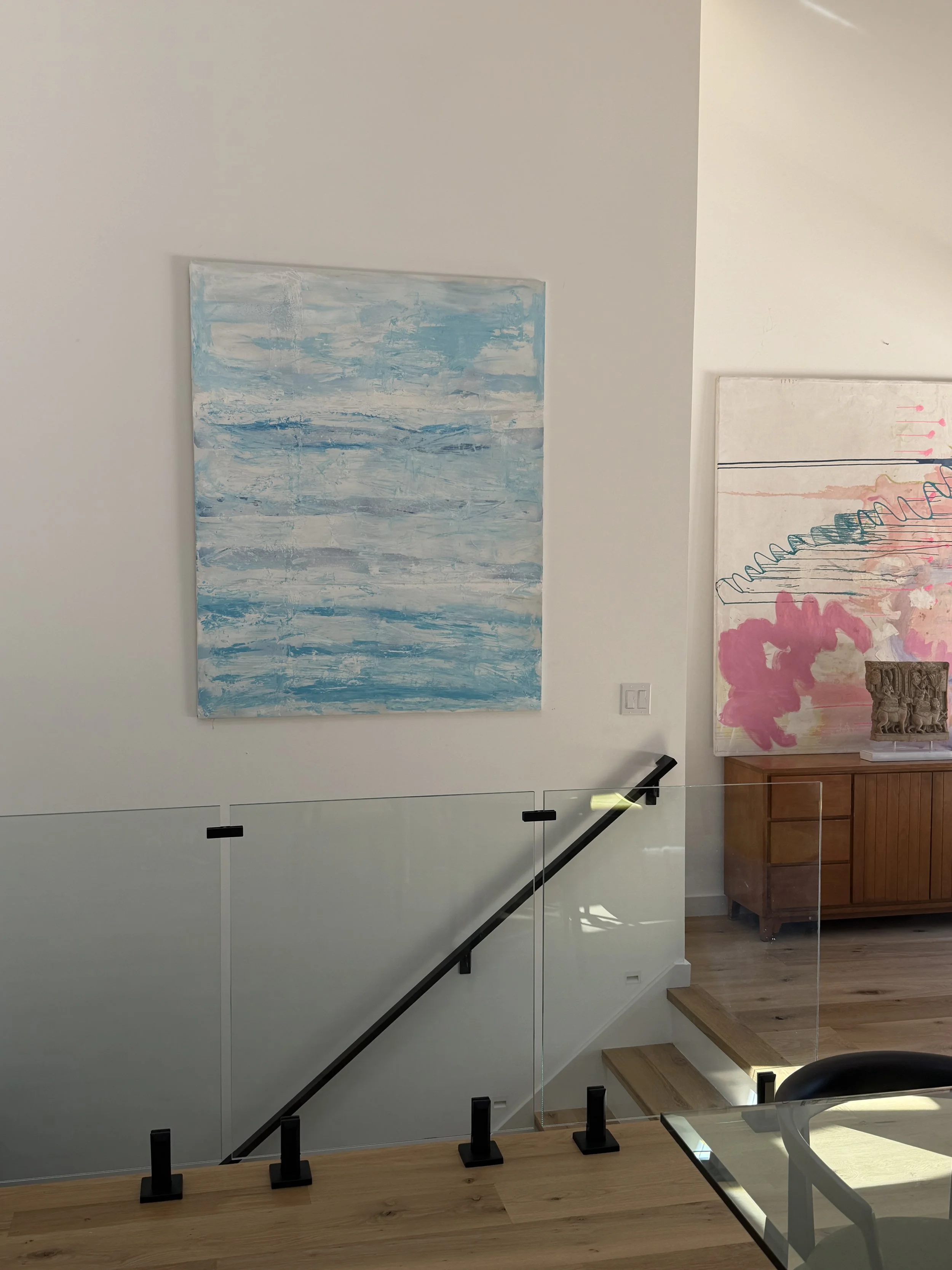Art gallery with two large abstract paintings on white walls, wooden staircase with black railings, glass barrier, wooden floor, and a wooden cabinet with a carved sculpture on top.