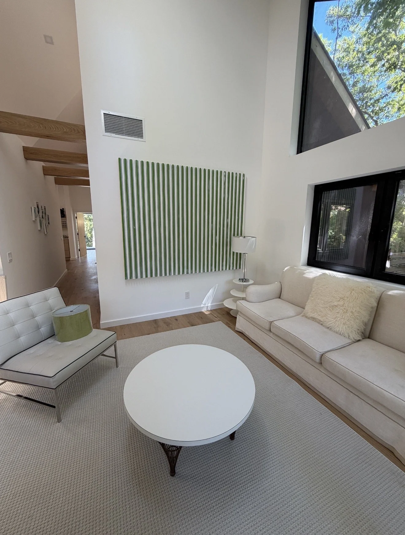 Living room with white furniture, including a sofa with a fur pillow and a matching chair with a green lampshade, a round white coffee table with a metal base, a white side table with a metal lamp, a large green and white striped wall art piece, hard