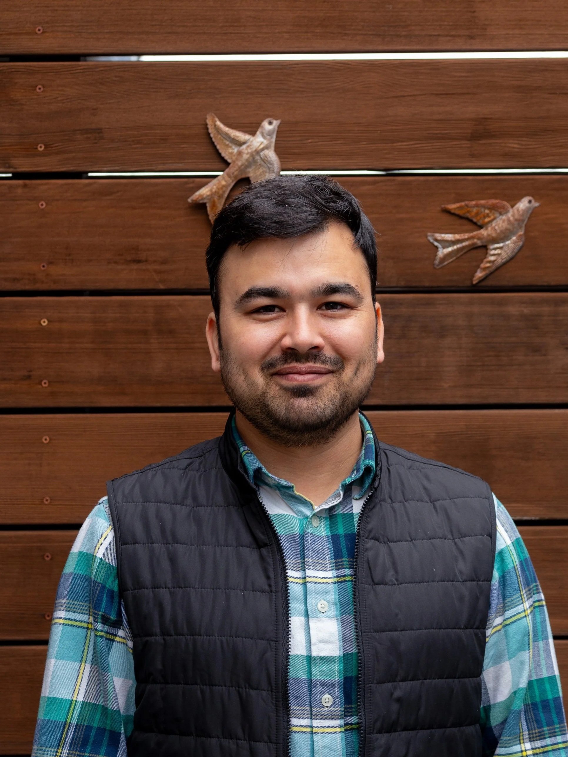 A man with dark hair and a beard smiling, standing in front of a wooden wall with metal bird decorations.