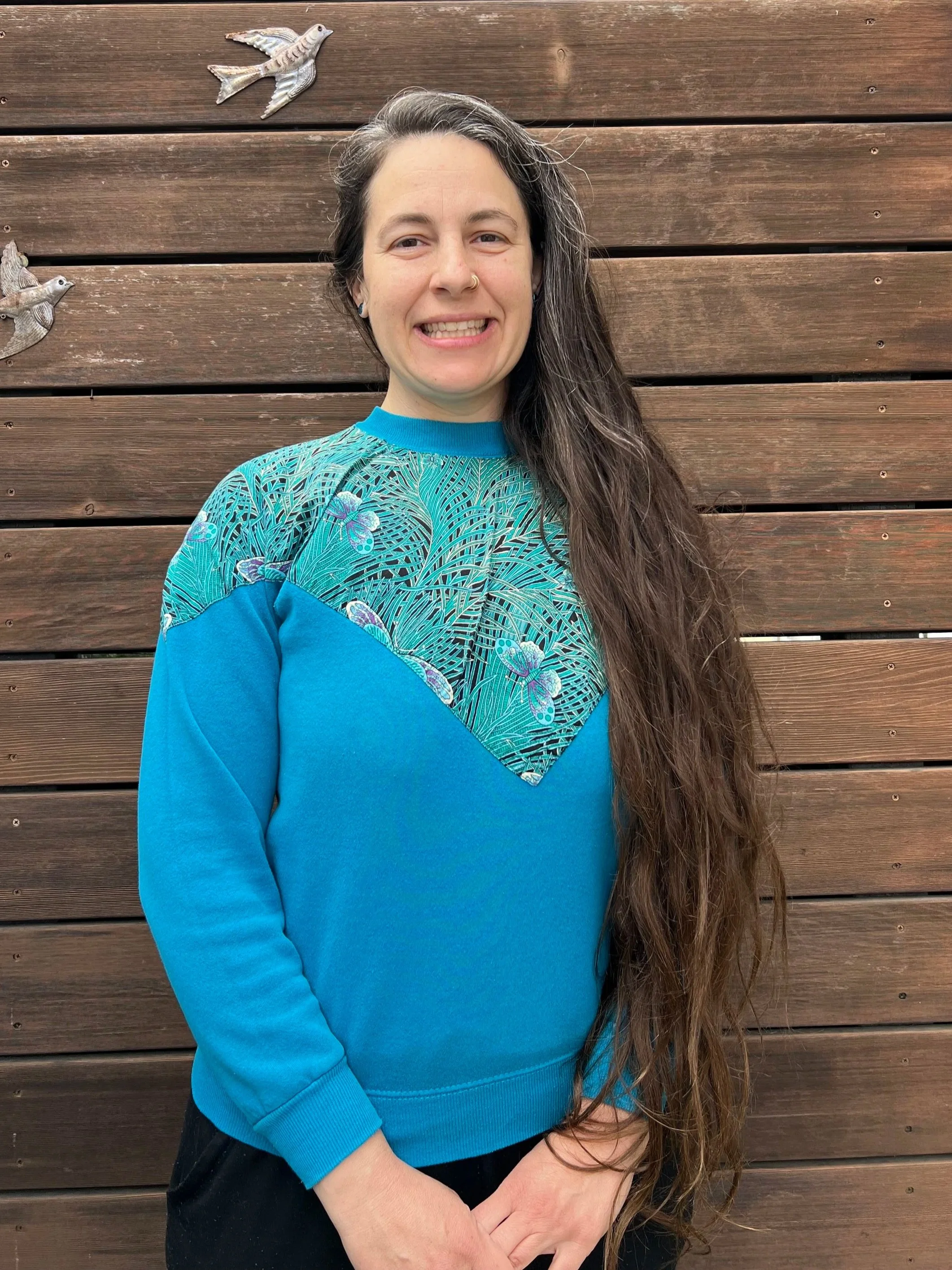 A woman with long brown hair, smiling, standing in front of a wooden wall decorated with metal bird sculptures.