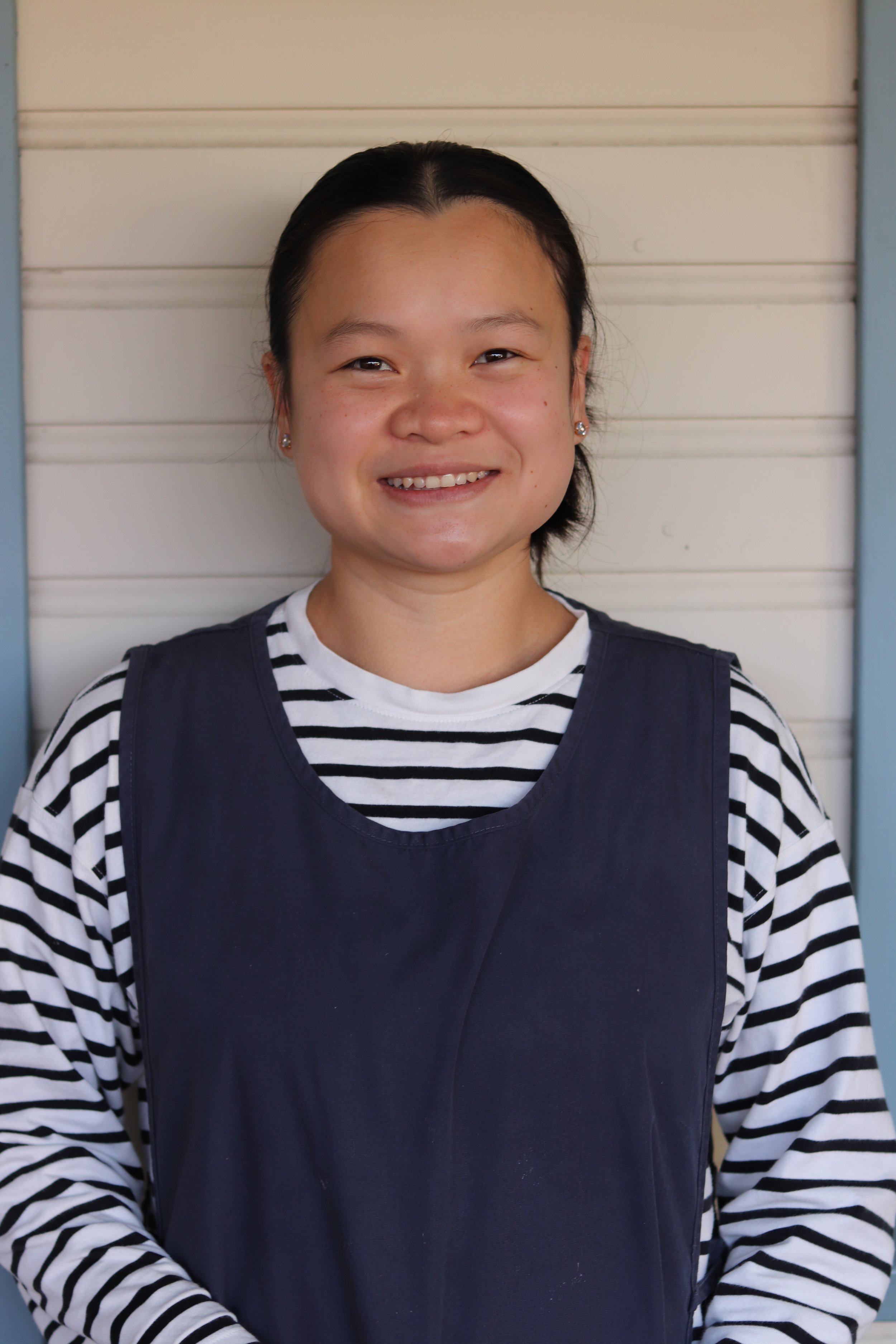 A young woman with dark hair tied back, wearing a blue t-shirt, smiling at the camera against a plain wall.