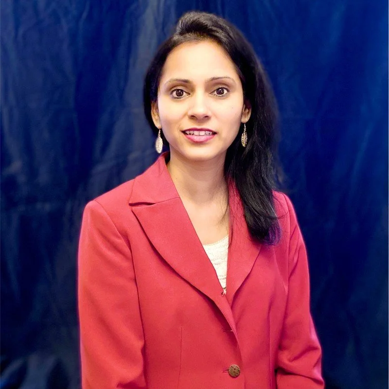 A woman with long black hair, wearing a red blazer and earrings, standing in front of a dark blue backdrop.