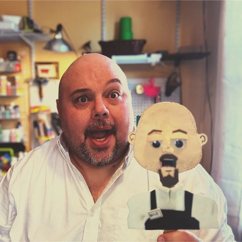 A man with a bald head and beard holding a cartoon-style paper cutout of himself. The man is smiling with an open mouth, in a workshop or craft room with shelves and tools in the background.