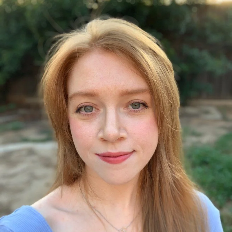 A young woman with light skin, light brown hair with reddish undertones, and blue eyes smiling outdoors with greenery in the background.