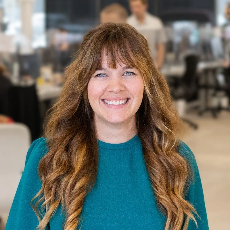 A woman with long wavy light brown hair and blue eyes smiling in an office environment.