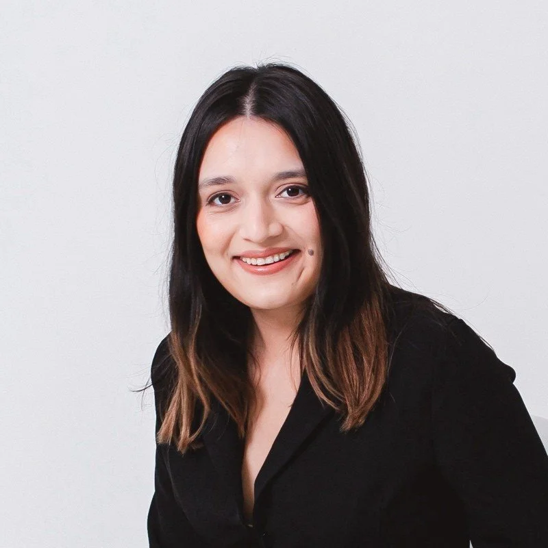 A woman with long dark hair, wearing a black shirt, smiling at the camera, against a plain white background.