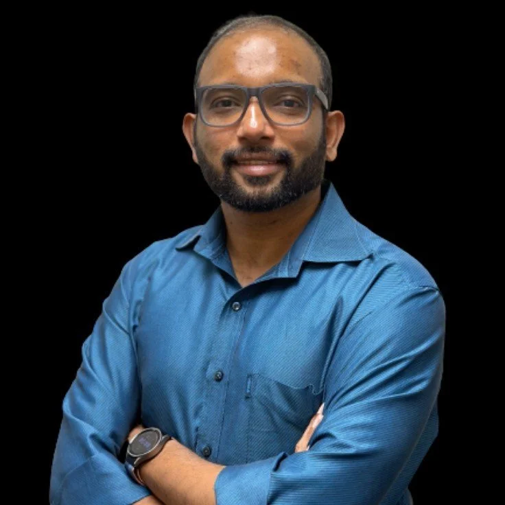 A man with short hair, beard, glasses, wearing a blue button-up shirt and a wristwatch, smiling with arms crossed against a black background.