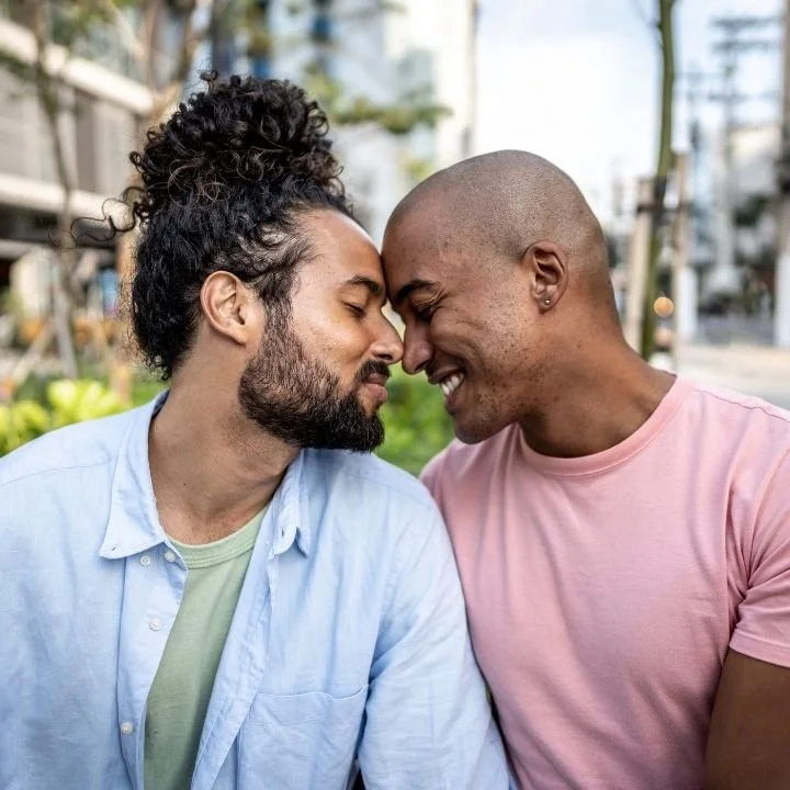 Two men with their foreheads touching, smiling with eyes closed, outdoors in a city setting.