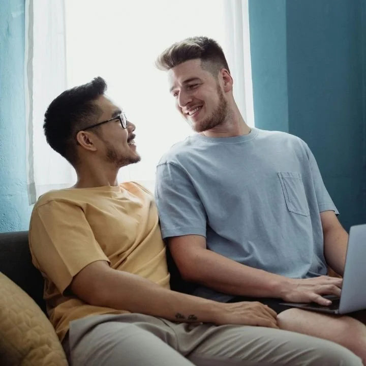 Two men sitting close together on a couch, smiling at each other, with a laptop open in front of them in a bright room.