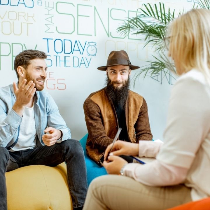 Two men and a woman having a conversation in a modern office or lounge area with a white wall and green plant in the background.