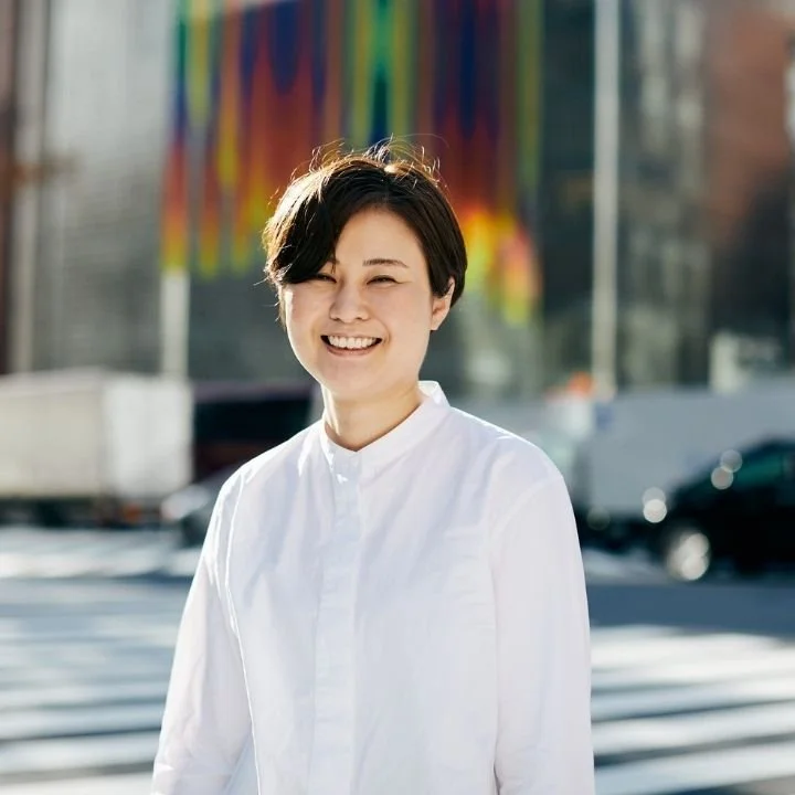 Young woman with short dark hair, smiling, wearing a white button-up shirt, standing outdoors in front of a colorful building and blurry cars.