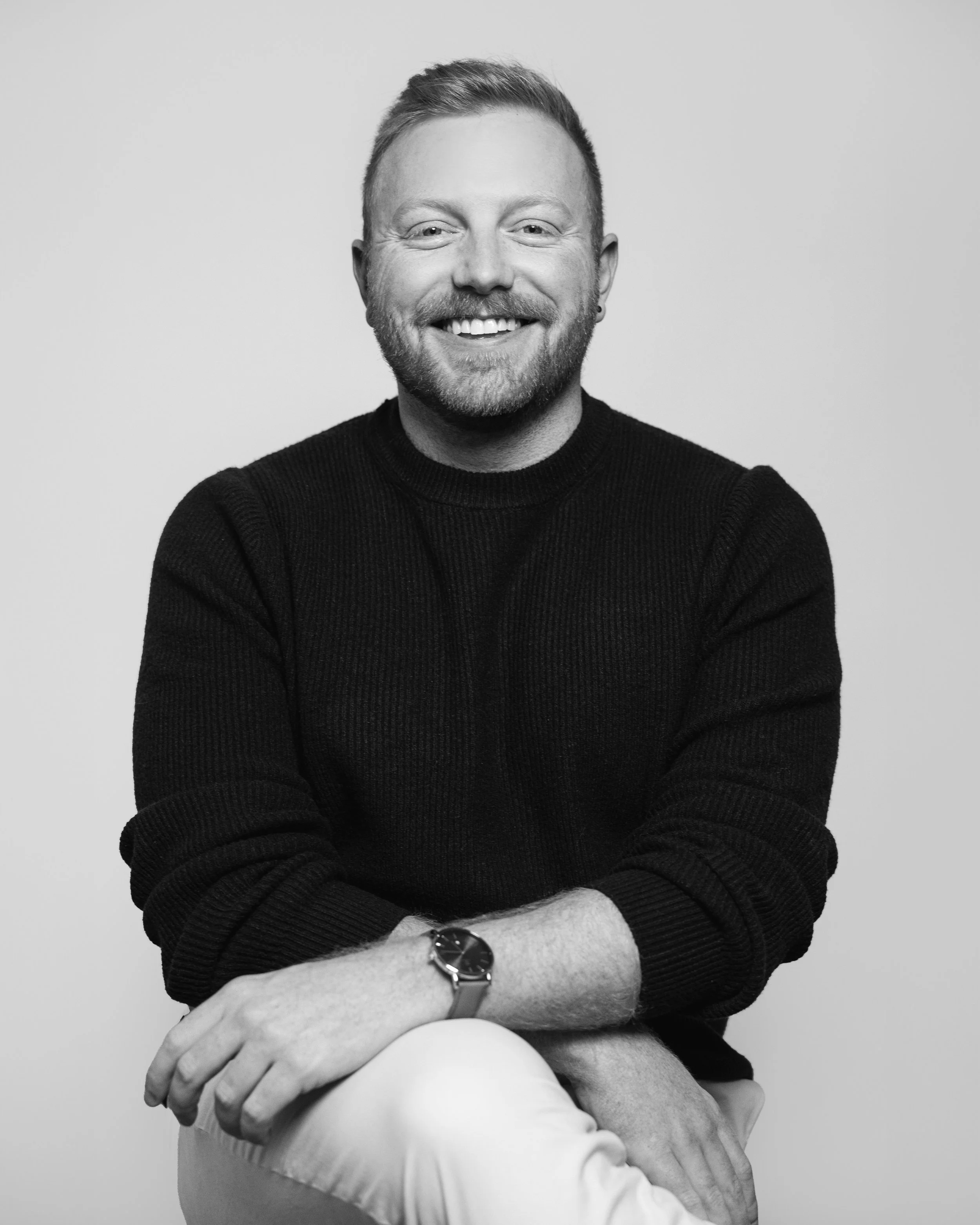 Black and white portrait of a smiling man with short hair, beard, wearing a dark sweater, sitting with arms crossed, on a plain light background.