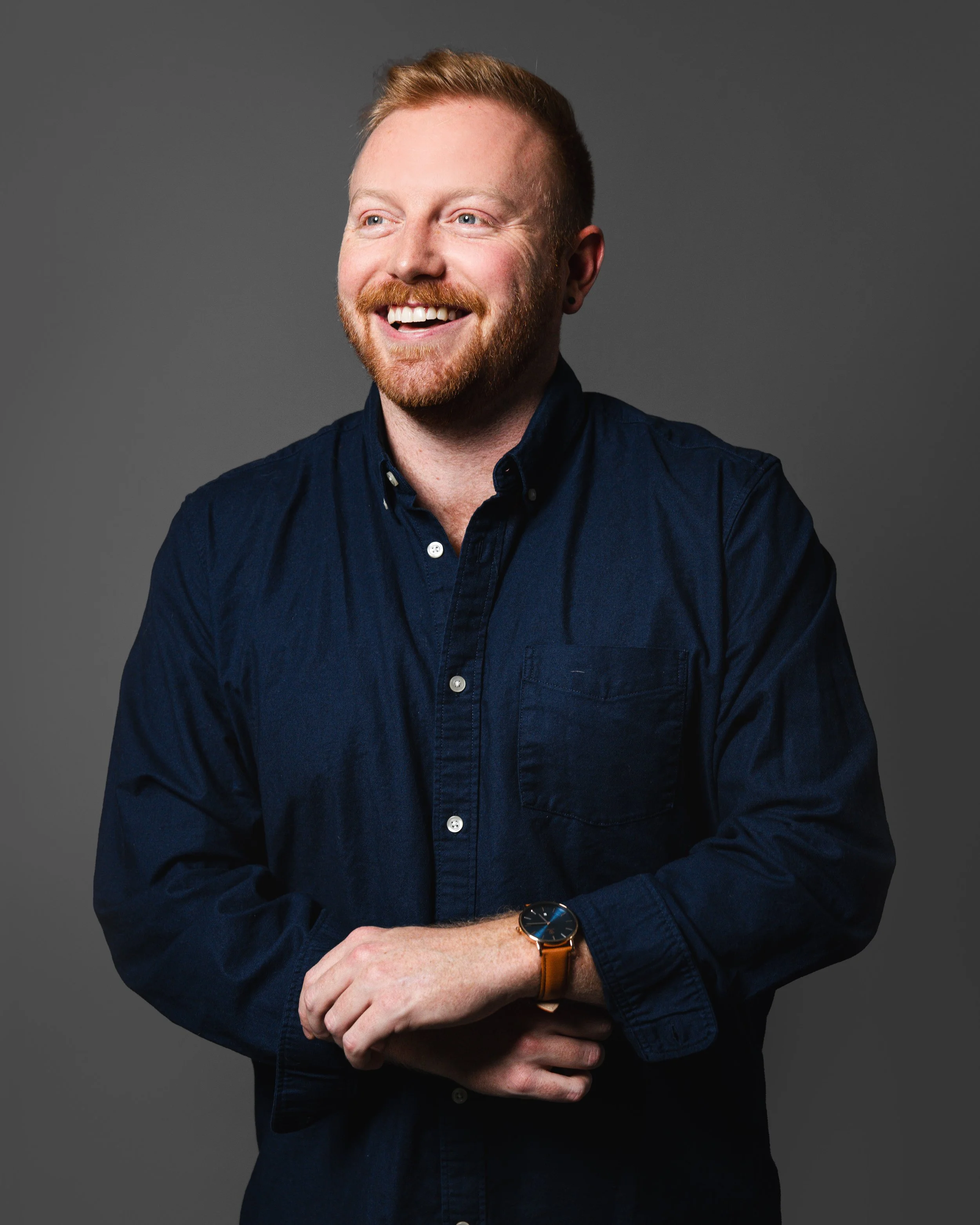 A smiling man with light skin, red hair, a beard, wearing a dark blue button-up shirt and a wristwatch, standing against a gray background.
