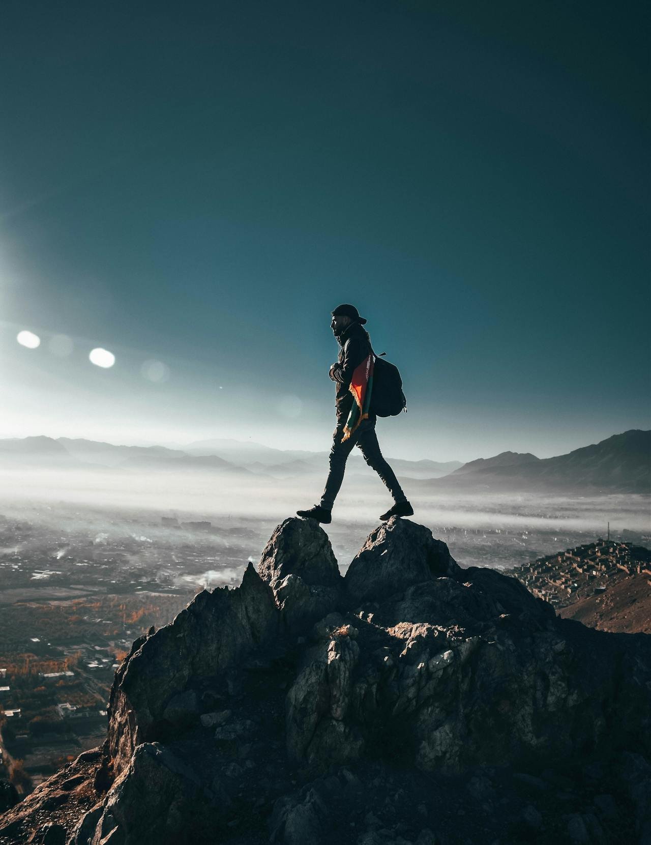 A person standing on a mountain rock formation with a backpack, overlooking a foggy landscape with mountains and a valley.
