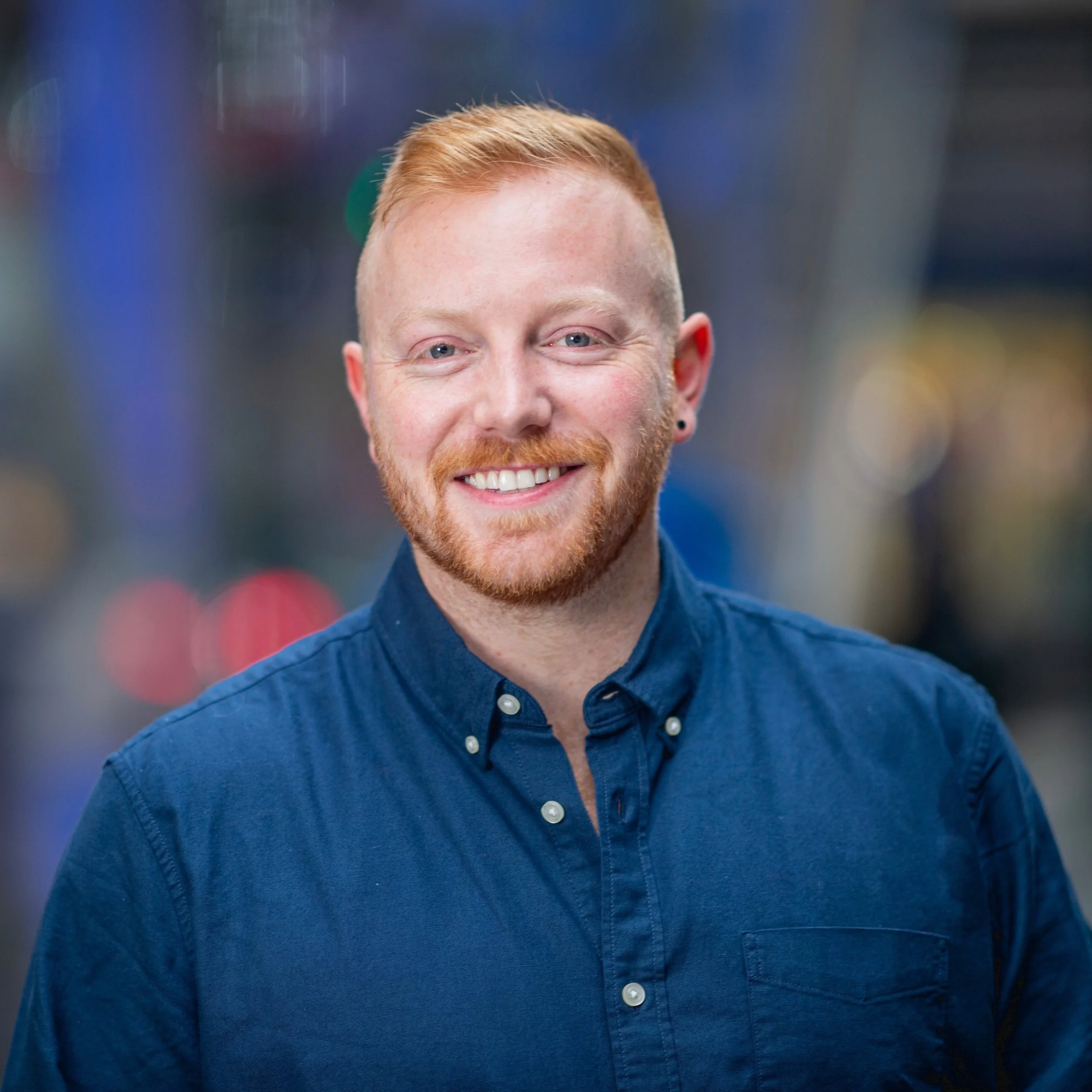 Headshot of a smiling man with red hair and beard, wearing a navy blue button-up shirt, in an urban setting at night.