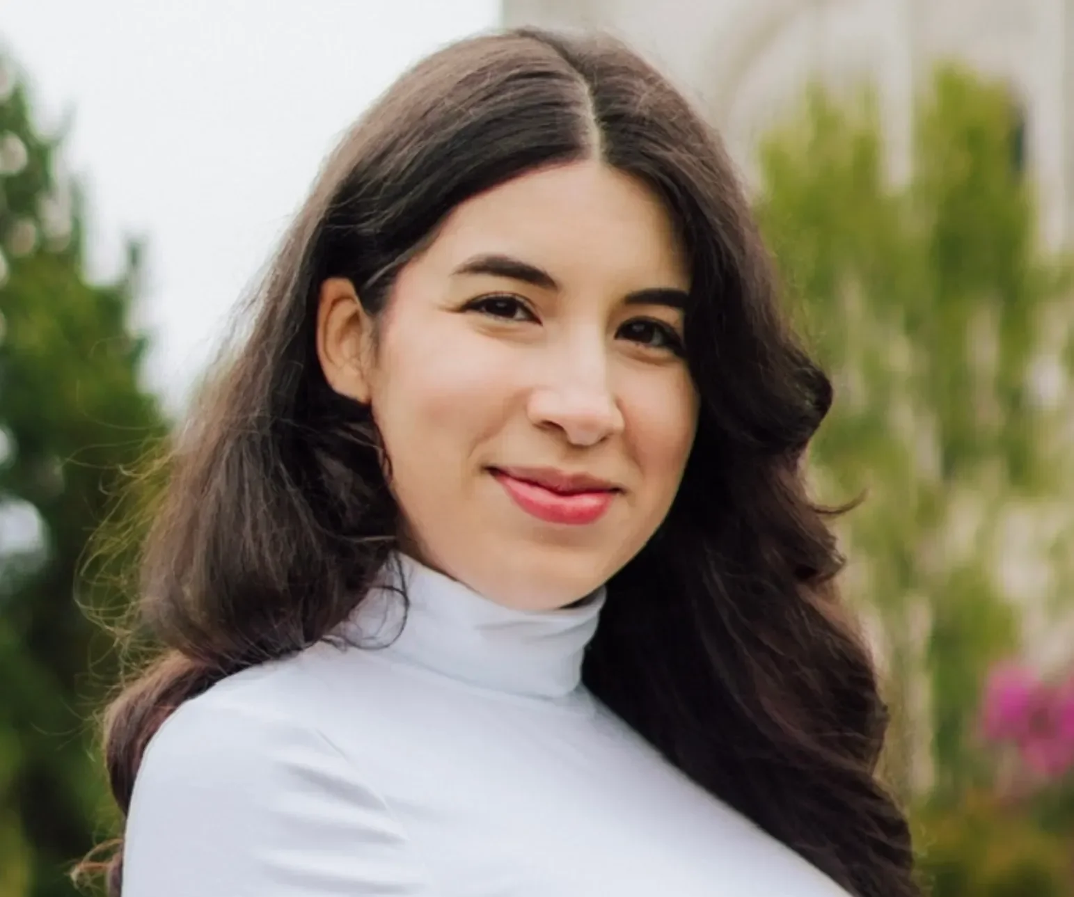 A woman with long dark hair wearing a white turtleneck standing outdoors, smiling with greenery and flowers in the background.