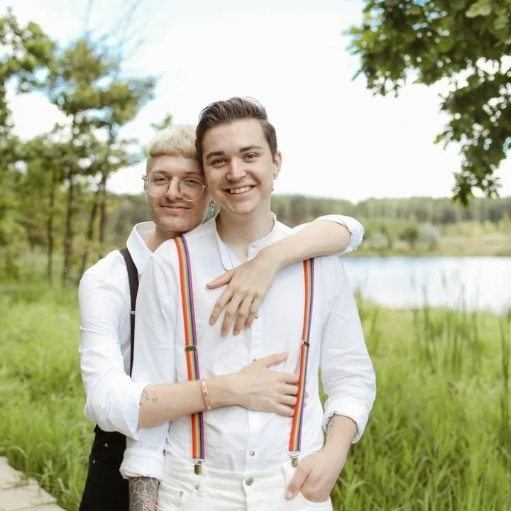 Two young men smiling, hugging outdoors near a lake with green trees and grass.