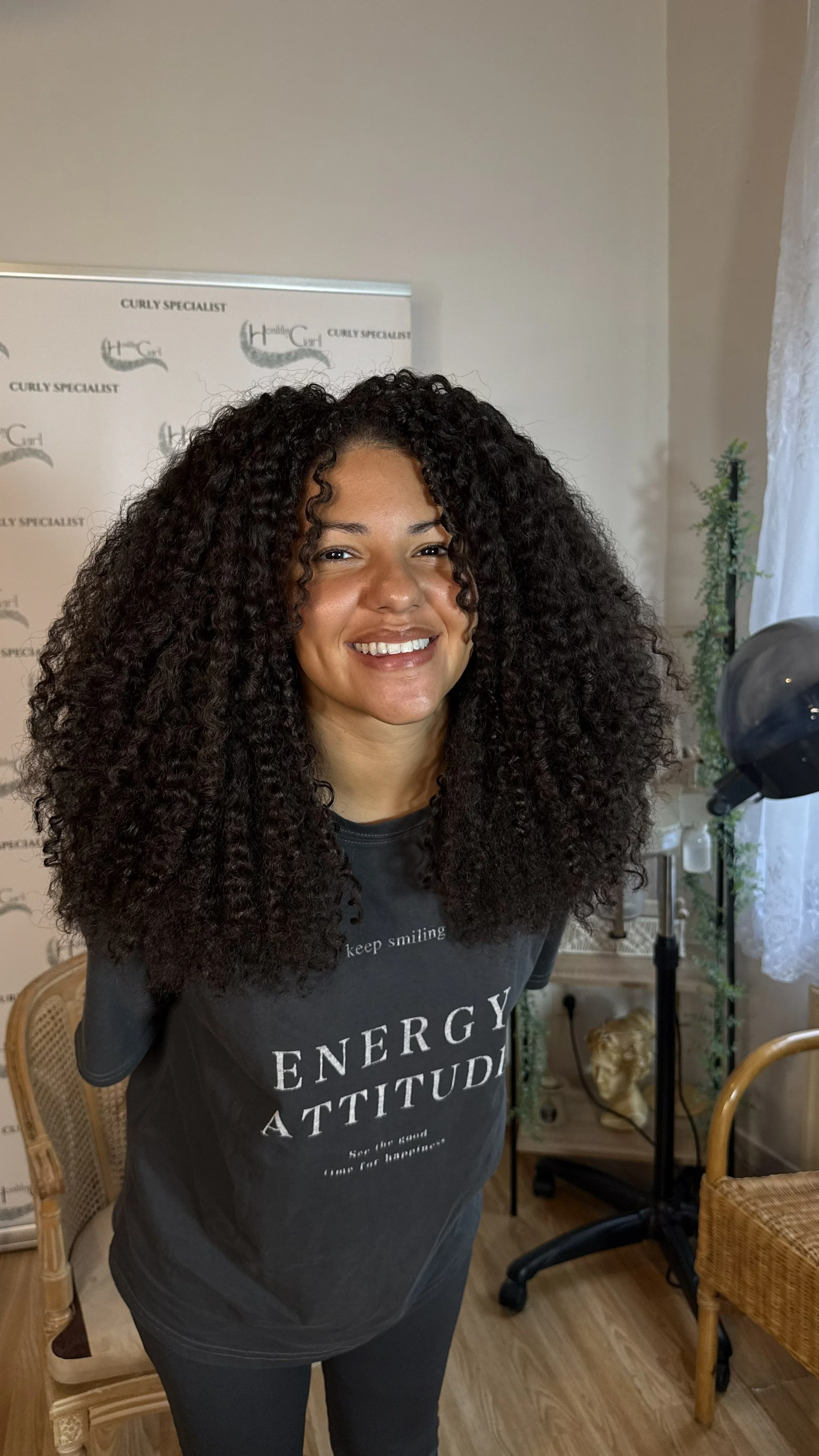 Une femme souriante avec des cheveux bouclés longs et noirs, portant un t-shirt noir avec écrit 'ENERGY ATTITUDE'. Elle se trouve dans une pièce avec un décor vintage et des plantes, près d'un chevalet de coiffure.