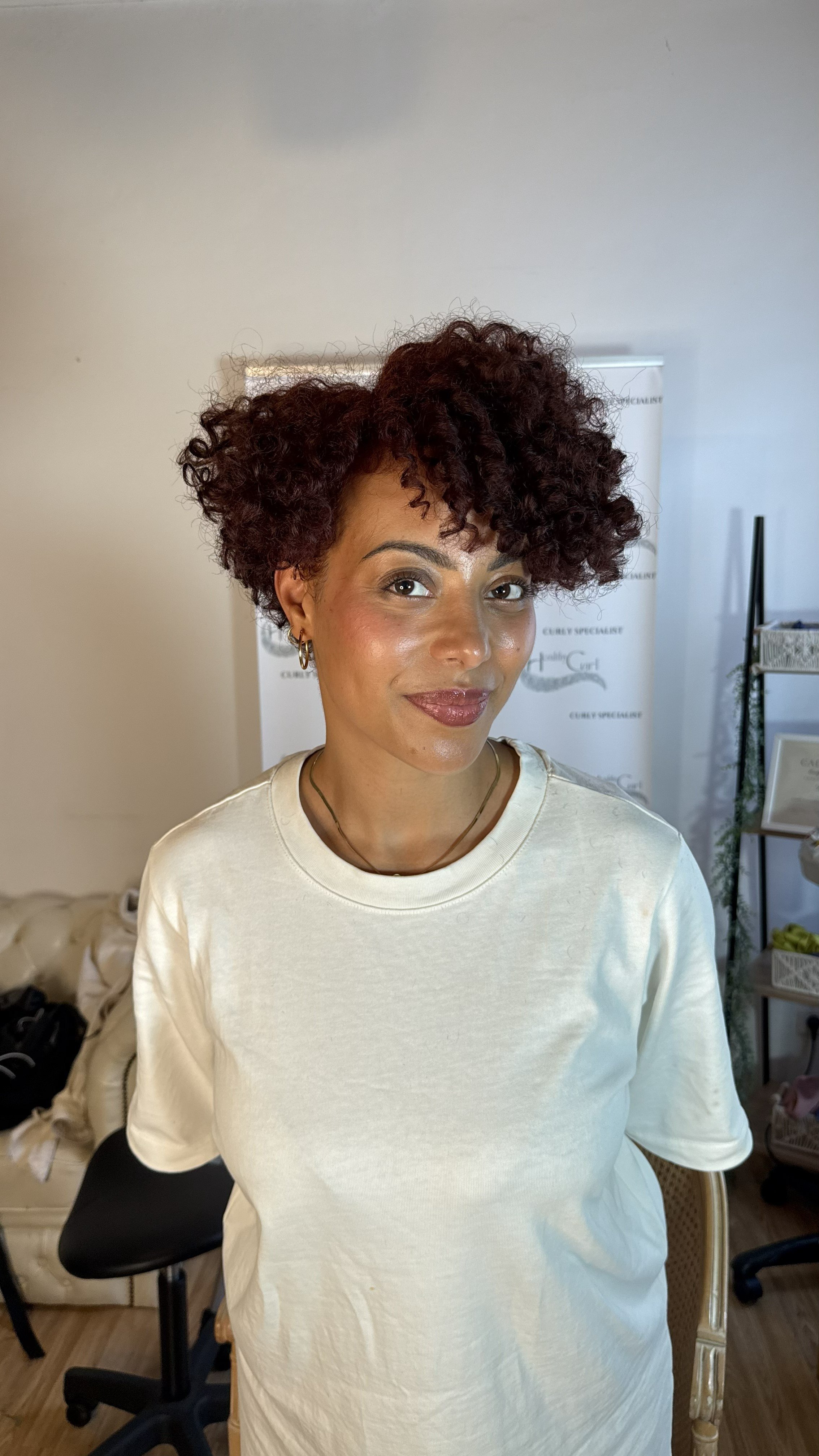 Une femme avec des cheveux bouclés et courts, portant un t-shirt blanc, sourit légèrement, dans un intérieur avec un fond blanc et des meubles.