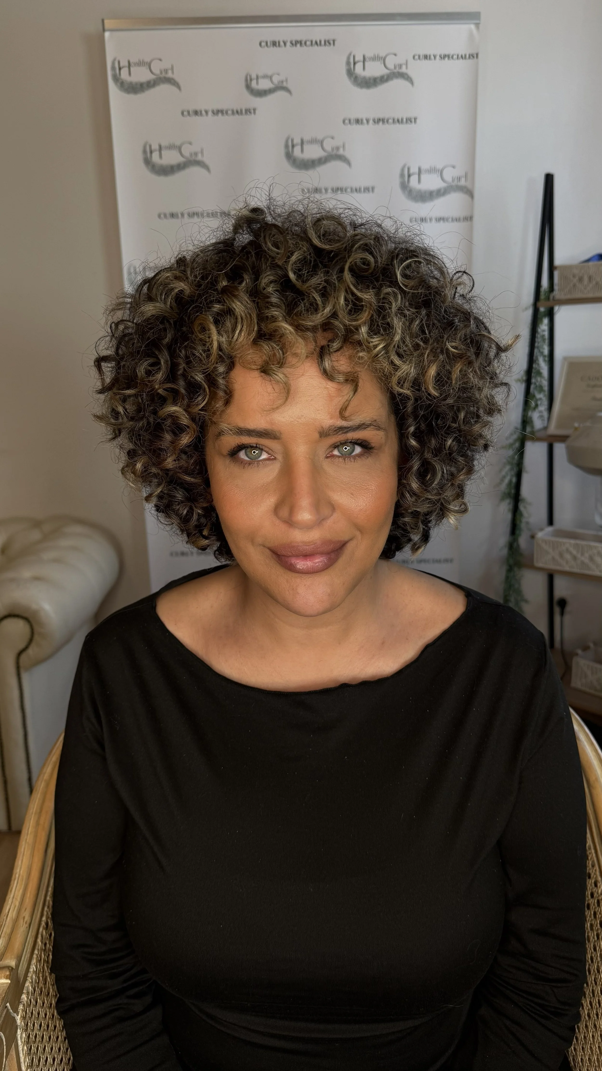 Portrait d'une femme aux cheveux bouclés et bruns, portant un haut noir, assise dans un fauteuil en osier, avec un fond de mur blanc et un tableau ou une affiche sur fond blanc avec le texte "Curly Specialist".