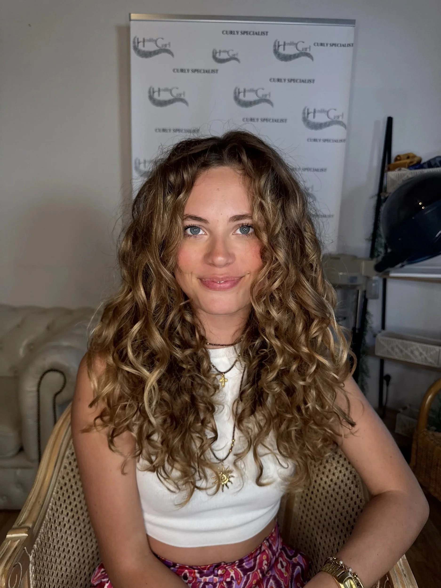 Jeune femme avec longs cheveux bouclés, portant un haut blanc, des bijoux dorés et un maquillage subtil, assise dans une pièce avec un mur gris et un poster "Curly Specialist" en arrière-plan.