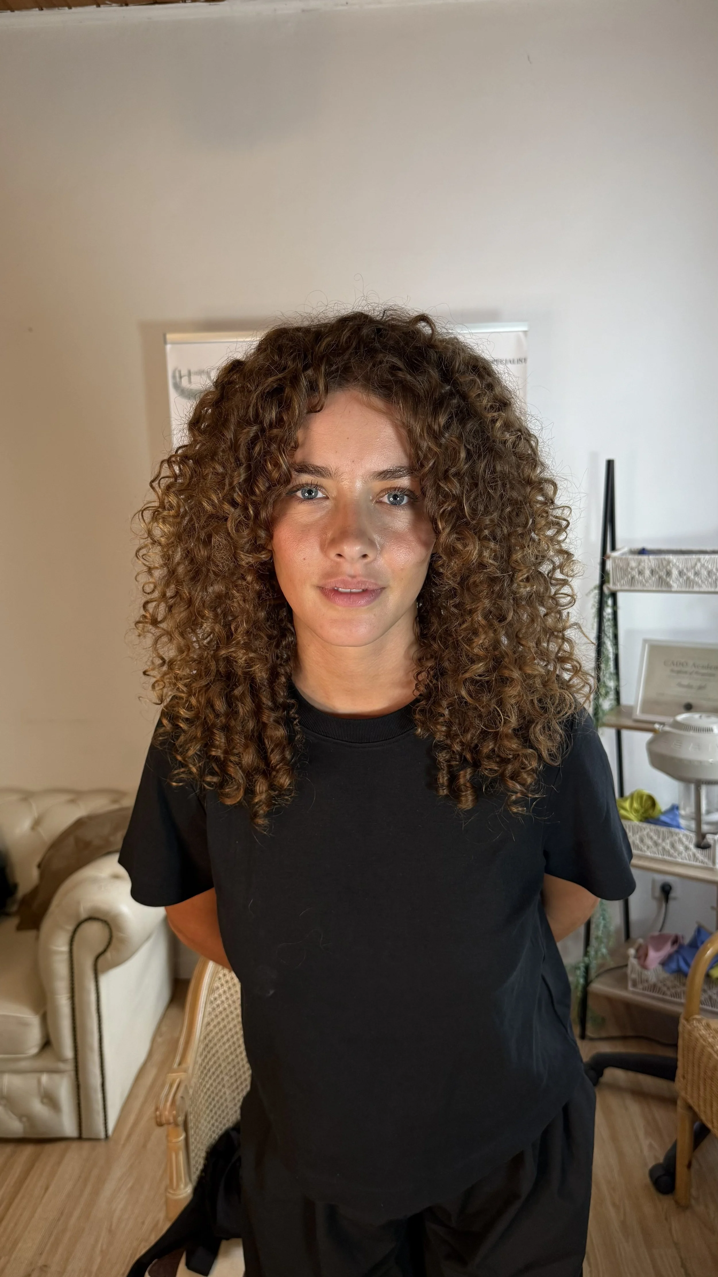 Une femme aux cheveux bouclés bruns, portant un t-shirt noir, se tient dans une pièce moderne avec des meubles en bois et une étagère en arrière-plan.