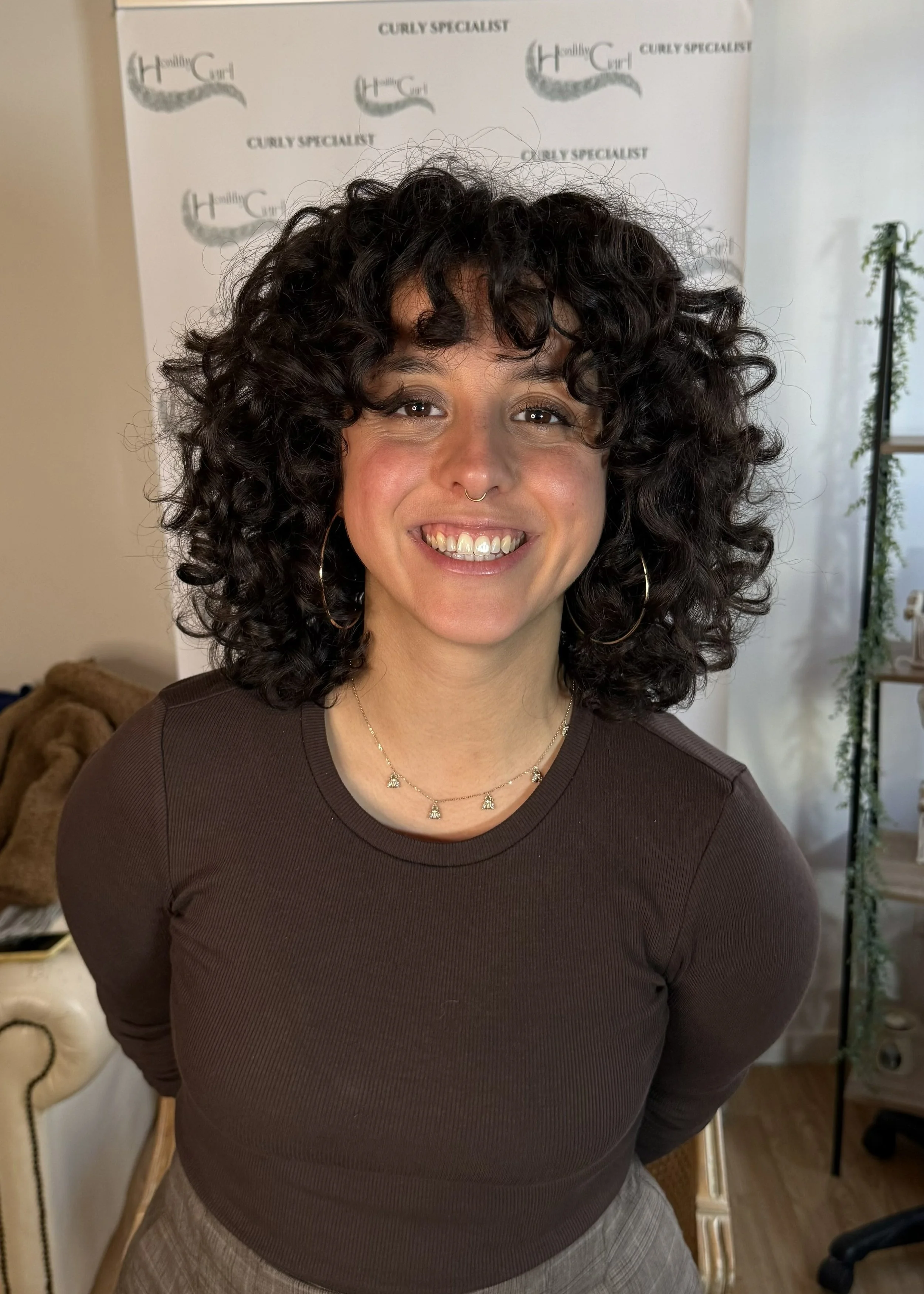 Portrait d'une femme avec des cheveux bouclés noirs, portant un collier et un sweat brun, souriant devant un fond blanc avec des logos.