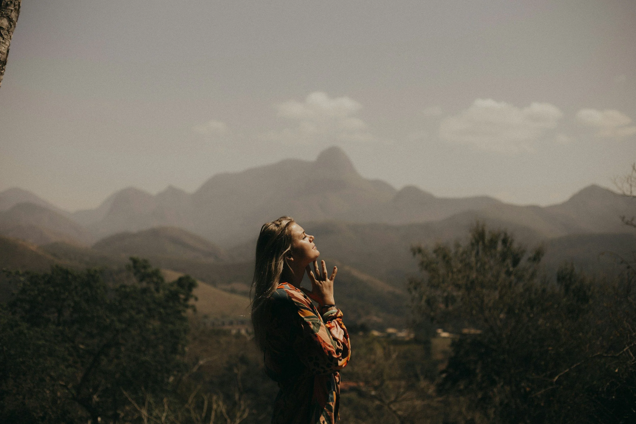 A woman with long hair stands outdoors in a landscape with mountains in the distance, eyes closed, head slightly tilted, and hand resting on her neck, dressed in a colorful jacket.