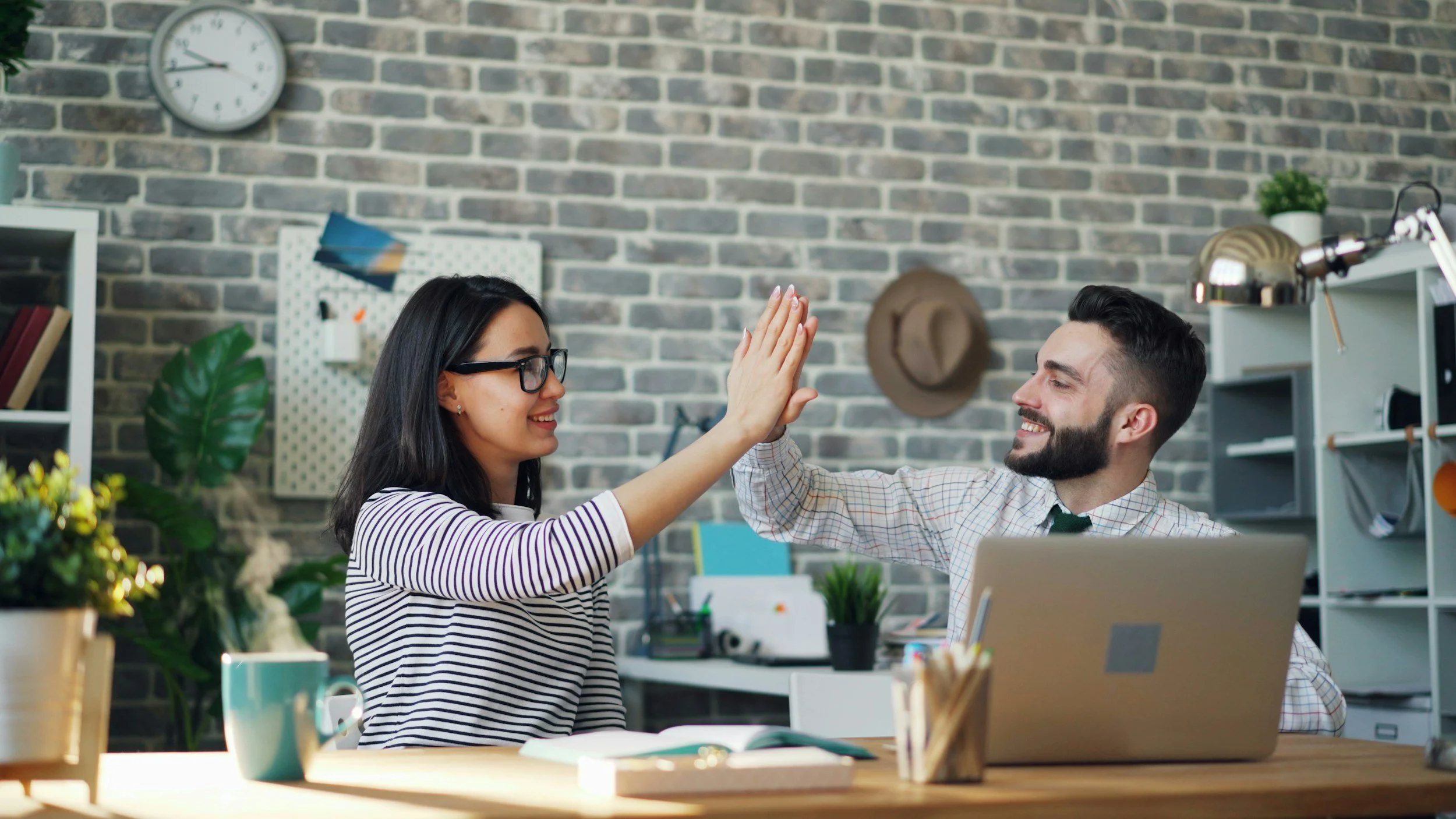 A woman and man in an office high-fiving each other, smiling. The woman is wearing glasses and a striped shirt, and the man has a beard and is wearing a checkered shirt. The desk has a laptop, notebooks, and a plant. The background features a brick wall, shelves, and office supplies.