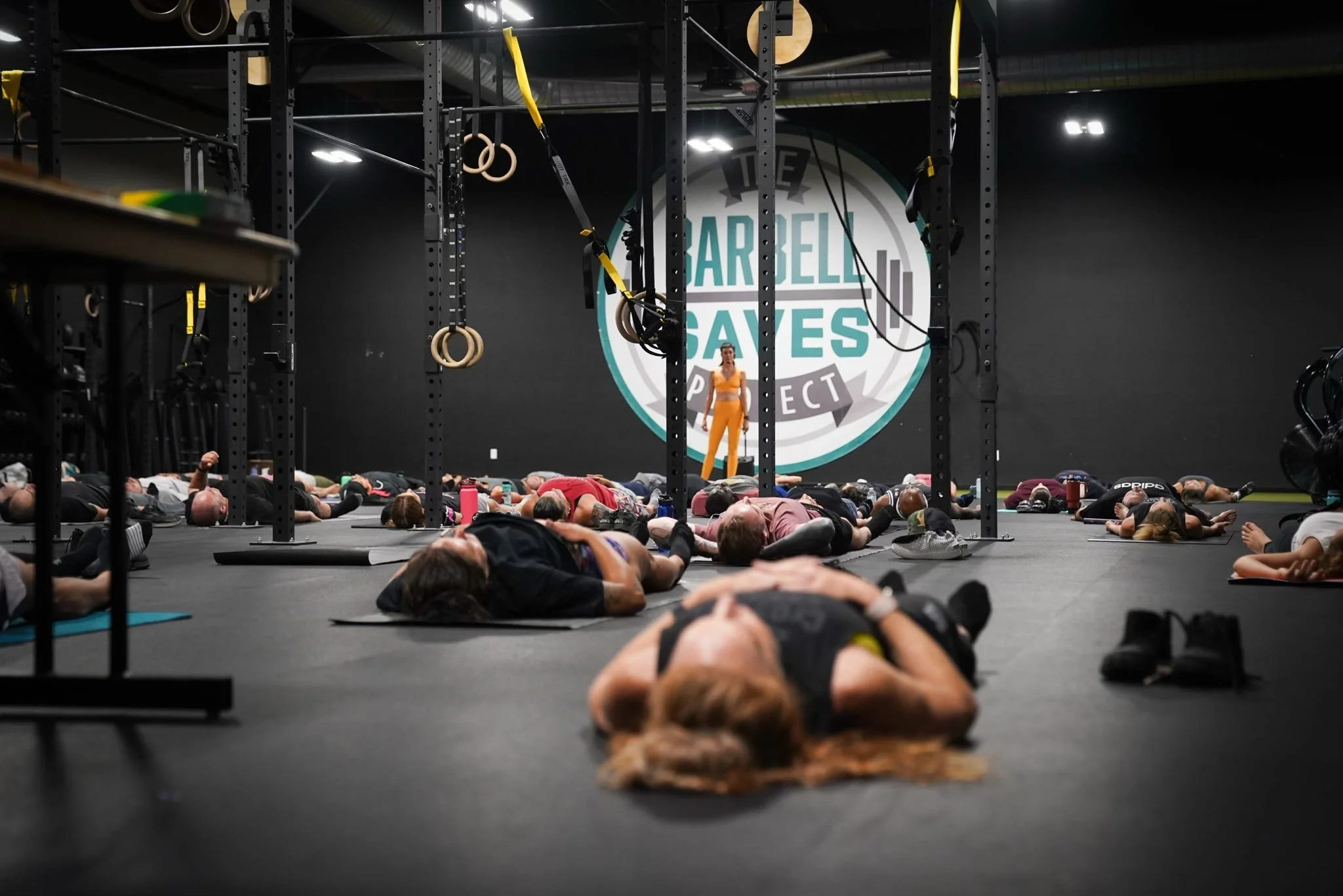 Group of people lying on the floor in a gym during a fitness class, with a trainer standing at the front near a wall with a large logo that reads 'The Barbell Saves Project'.